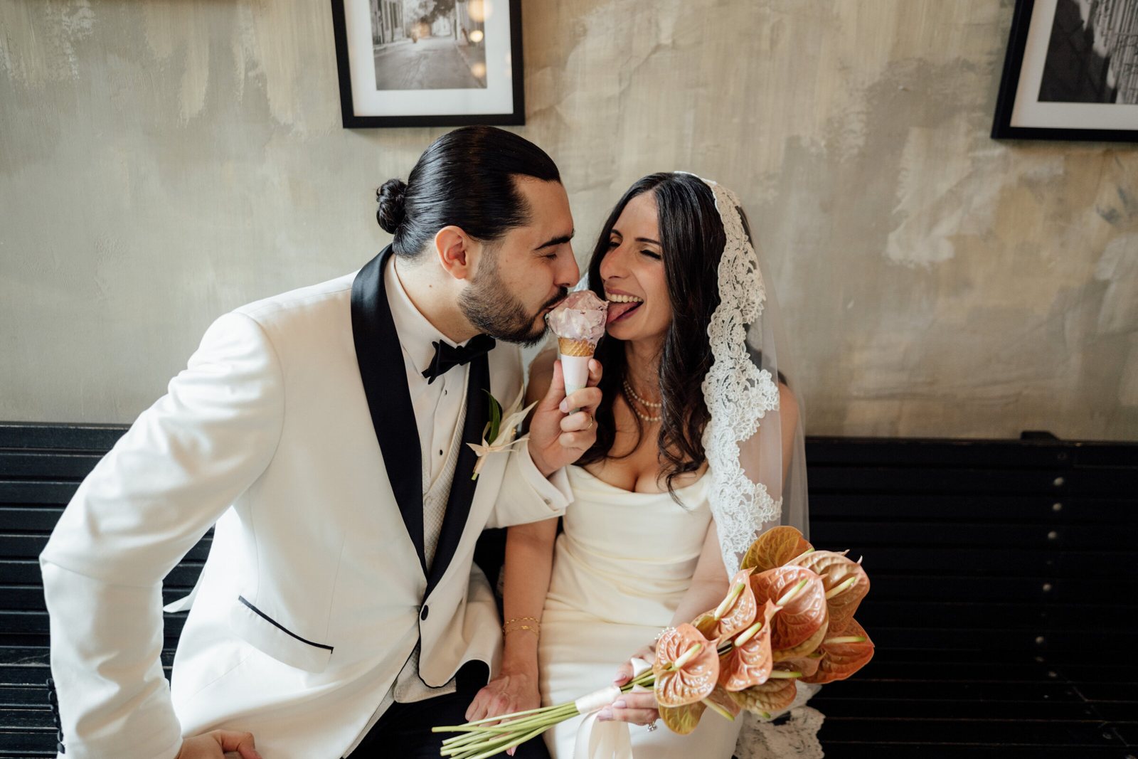A groom in a white tuxedo jacket laughing as his bride takes a bite of his gelato
