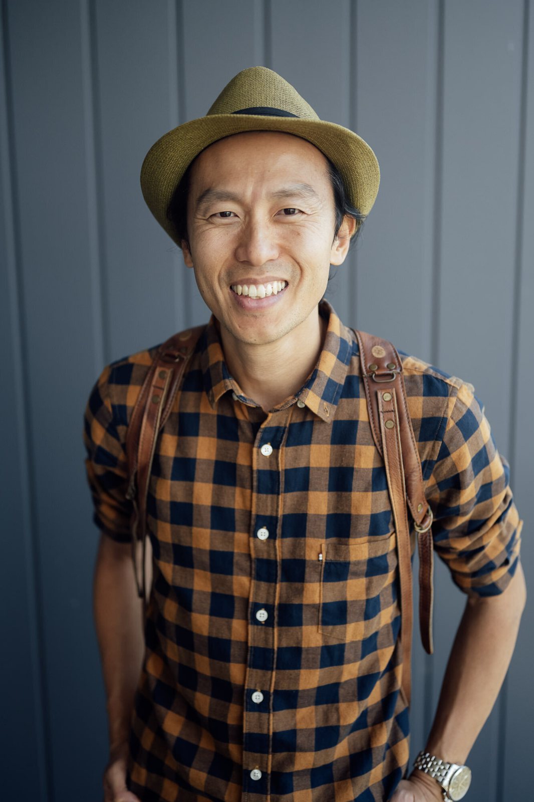 Kevin smiling in a tan hat and checked shirt against a grey wall, camera strap over his shoulder