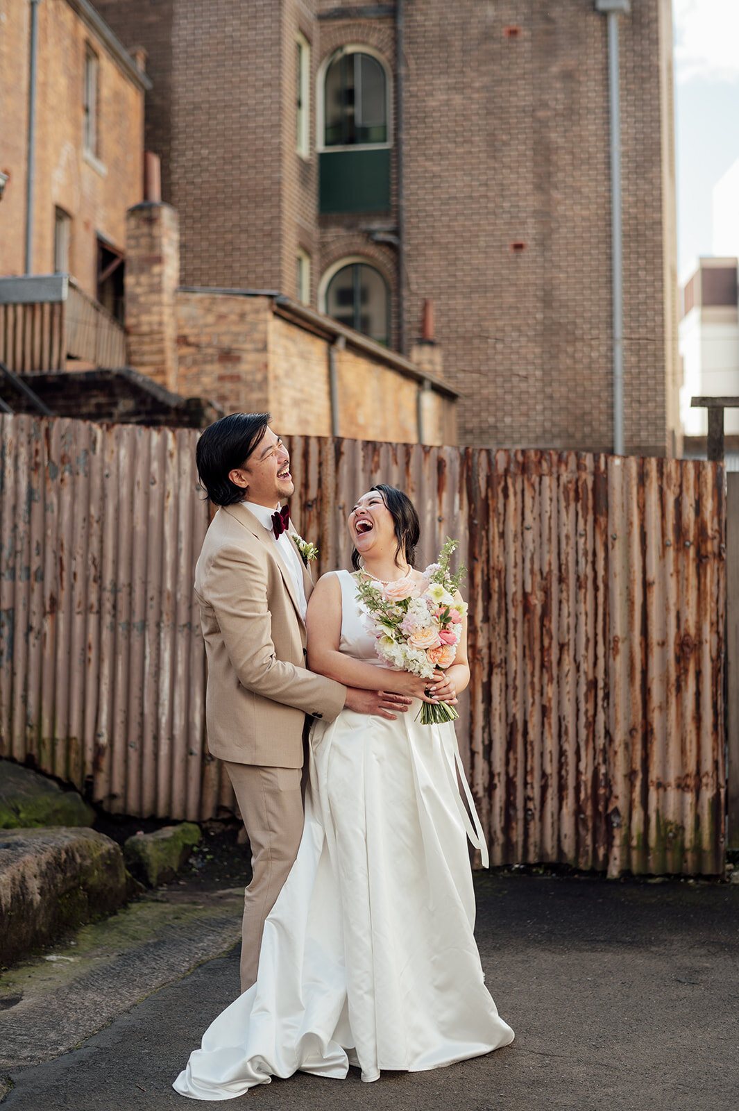 A bride laughing in her groom&rsquo;s arms in a city laneway beside a brick building