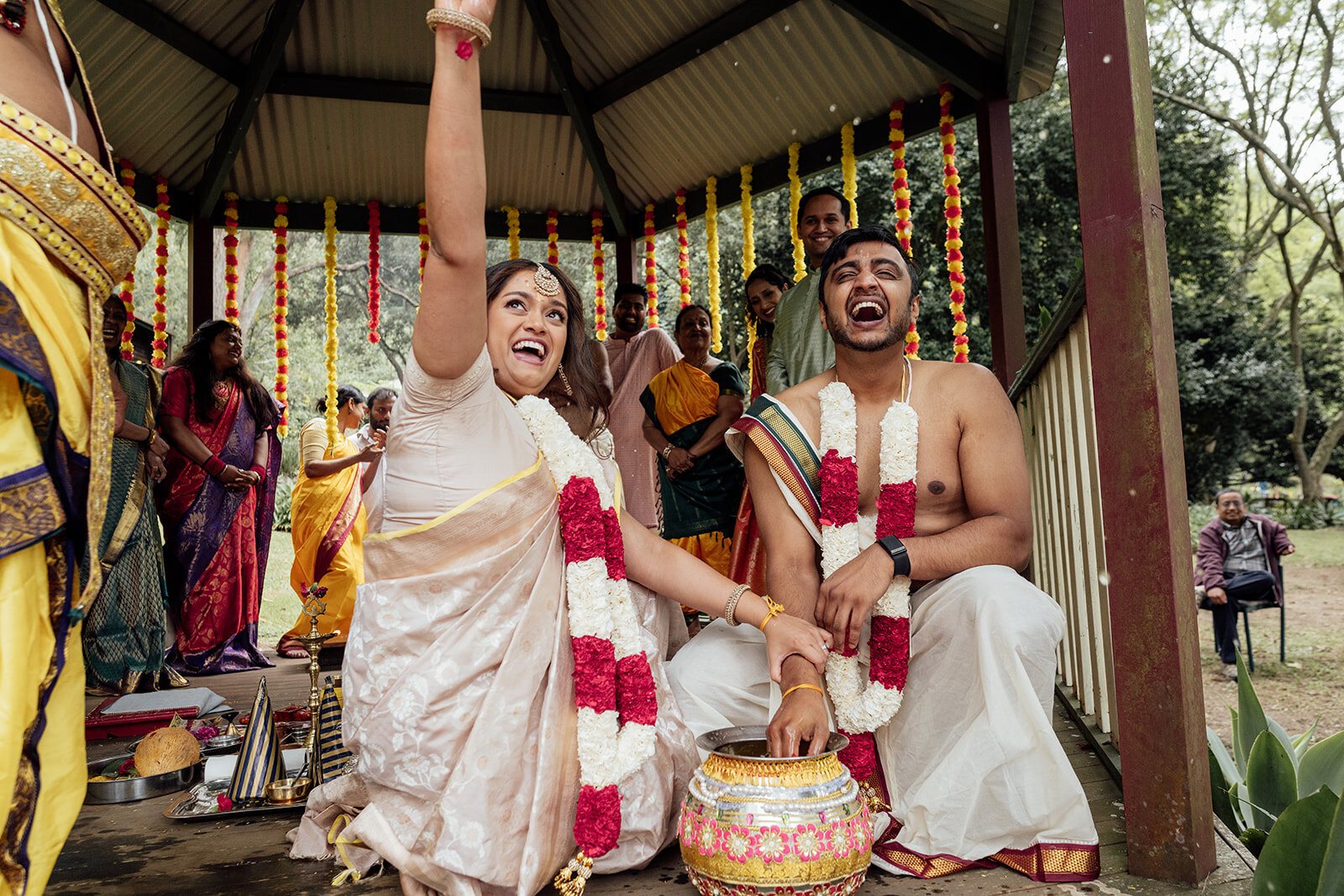 A bride throwing her arm up and laughing during a Tamil ceremony under marigold garlands