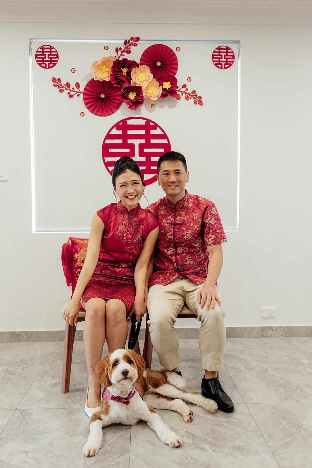 A couple in red at their tea ceremony, sitting under a double happiness sign with their dog at their feet